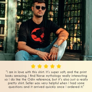 May include: Black t-shirt featuring a red graphic of a raven in front of a red circle. The shirt is worn by a person wearing sunglasses and a watch. The shirt design may be inspired by Norse mythology.