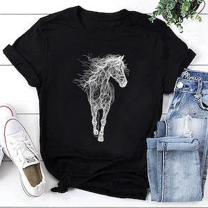 May include: Black t-shirt featuring a white, stylized illustration of a running horse. The horse design has a flowing, ethereal quality. The shirt is paired with blue jeans and white sneakers, suggesting casual wear.
