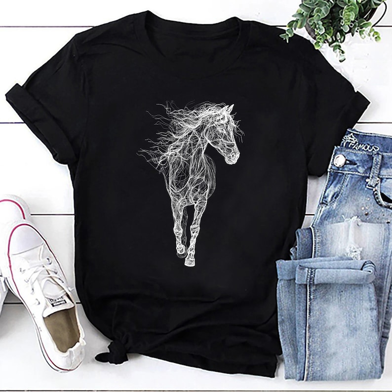 May include: Black t-shirt featuring a white, stylized illustration of a running horse. The horse design has a flowing, ethereal quality. The shirt is paired with blue jeans and white sneakers, suggesting casual wear.