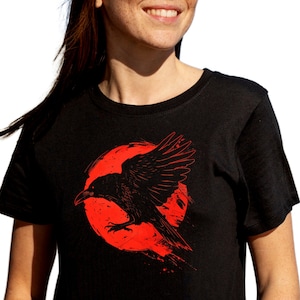 May include: Black t-shirt featuring a red graphic of a raven in flight against a red circular background. The raven is detailed with visible feathers and a sharp beak. The design has a distressed, artistic style.
