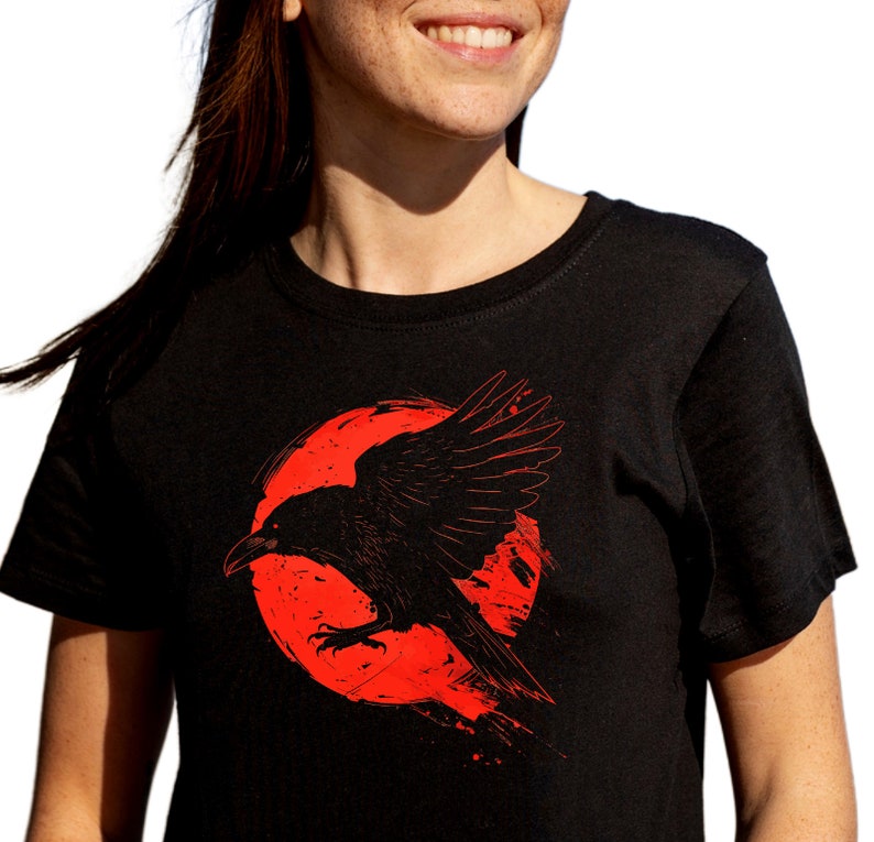May include: Black t-shirt featuring a red graphic of a raven in flight against a red circular background. The raven is detailed with visible feathers and a sharp beak. The design has a distressed, artistic style.
