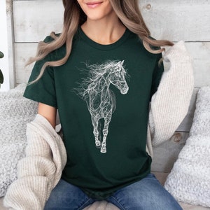 May include: Dark green t-shirt featuring a white, artistic illustration of a horse. The horse design appears to be made of interconnected lines, giving it a unique, ethereal look. The shirt is a crew neck style and appears to be made of soft material.