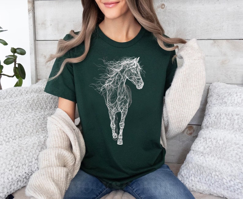 May include: Dark green t-shirt featuring a white, artistic illustration of a horse. The horse design appears to be made of interconnected lines, giving it a unique, ethereal look. The shirt is a crew neck style and appears to be made of soft material.