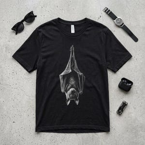 May include: Black t-shirt with a detailed grayscale illustration of a bat hanging upside down. The bat's wings are spread, and its face is visible. The shirt has a crew neck.