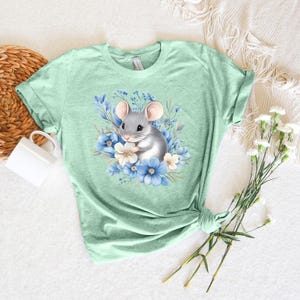 May include: A light green t-shirt with a gray mouse surrounded by blue and white flowers.