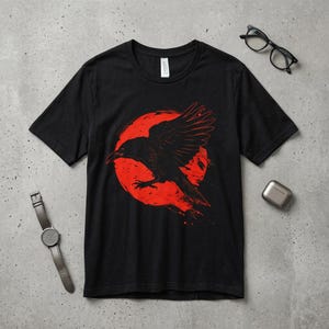 May include: Black t-shirt featuring a graphic of a black raven in flight against a red circle. The raven's wings are spread, and the design has a distressed, vintage look. The shirt is a crew neck style.