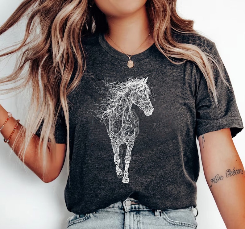 May include: Dark gray t-shirt featuring a white, artistic outline of a running horse. The design has a flowing, ethereal quality, with the horse's mane and tail appearing to blend into the background. The shirt has short sleeves and a crew neck.