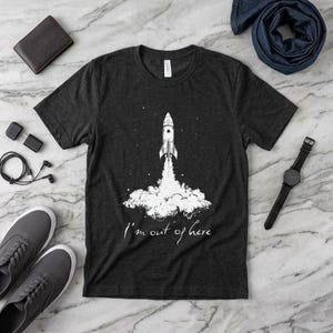 Może przedstawiać: Ciemnoszara koszulka z białą grafiką rakiety startującej, otoczonej gwiazdami i chmurami. Tekst "I'm out of here" jest napisany poniżej. Koszulka jest prezentowana na marmurowej powierzchni.