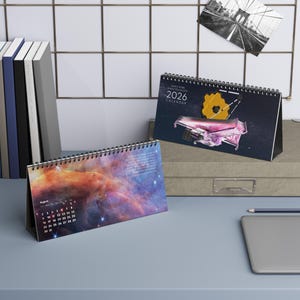 May include: Two spiral-bound desk calendars. One calendar features a nebula image, the other displays the James Webb Space Telescope with the text "2026 Calendar". The calendars are on a desk with a laptop and a pencil.