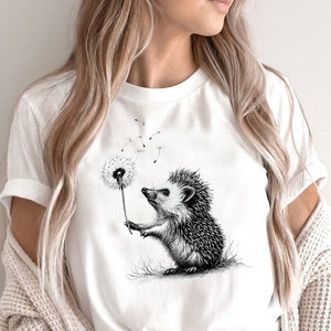 May include: A white t-shirt with a black and white illustration of a hedgehog blowing on a dandelion.