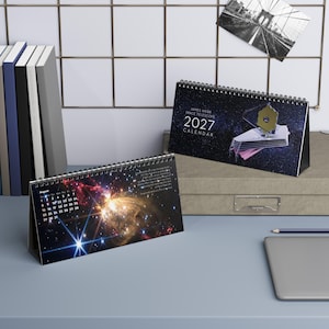 May include: A 2027 James Webb Space Telescope desk calendar featuring a dark blue space background with a telescope illustration on the cover and a vibrant nebula image on the open page, displaying the month of August with a grid of dates.