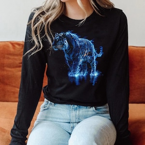 Puede incluir: Camiseta negra de manga larga con un diseño de tigre azul. El tigre tiene un efecto eléctrico y brillante, de pie en el agua. La camiseta está hecha de un tejido suave.
