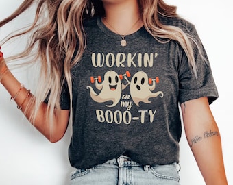 Puede incluir: Una camiseta gris oscuro con un gráfico de dos fantasmas levantando pesas. El texto en la camiseta dice "Workin' on my Boo-ty".