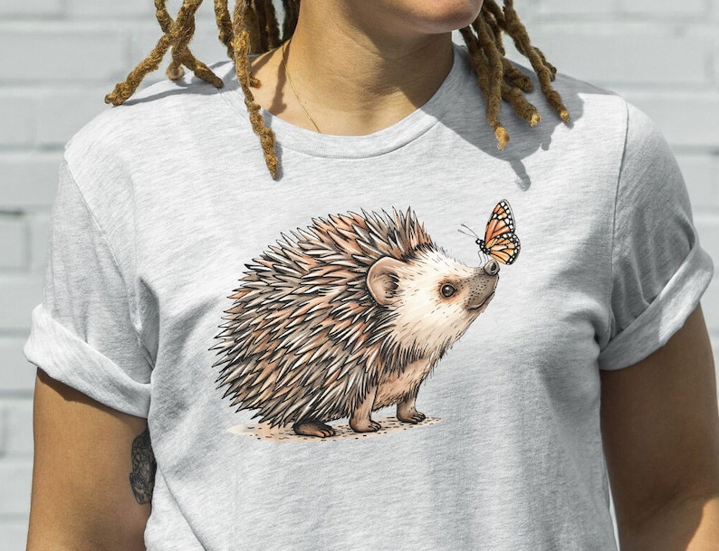 May include: A gray t-shirt with a brown and white hedgehog and a butterfly illustration. The hedgehog is looking at the butterfly.