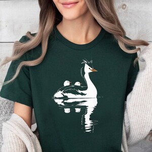Op de afbeelding: Donkergroen T-shirt met een witte illustratie van een vogel en twee kuikens op het water. De vogel heeft een lange nek en een kuif. Het ontwerp is eenvoudig en elegant, met een weerspiegeling in het water.