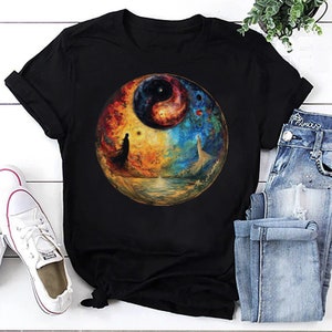 May include: Black t-shirt featuring a colourful yin yang symbol and a galaxy design. The symbol is surrounded by a circle of planets and stars.