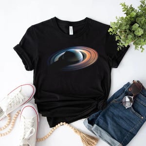 Może przedstawiać: Czarny T-shirt z grafiką przedstawiającą planetę z pierścieniami. Planeta jest ciemnoniebieska i fioletowa z jasnobrązowym pierścieniem wokół niej. T-shirt leży na białym tle z parą białych butów sportowych, niebieskimi dżinsowymi spodenkami i okularami przeciwsłonecznymi.