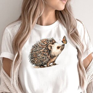 May include: White t-shirt with a brown and white hedgehog illustration and a small orange butterfly on the front.
