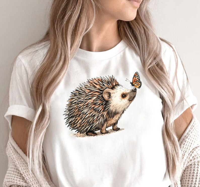 May include: White t-shirt with a brown and white hedgehog illustration and a small orange butterfly on the front.