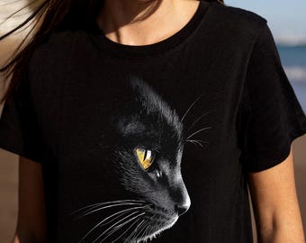 May include: Black t-shirt featuring a detailed graphic of a black cat's profile. The cat's eye is a bright yellow, contrasting with the dark fur. The design is centered on the shirt, creating a striking visual.