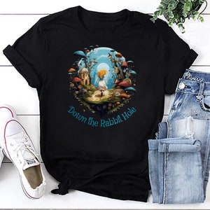 May include: Black t-shirt featuring a whimsical illustration of a girl walking towards a rabbit hole, surrounded by mushrooms and fantastical buildings. The text "Down the Rabbit Hole" is written in a teal color. The shirt is paired with blue jeans and white sneakers.