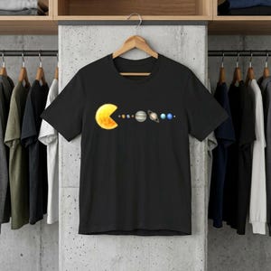 Op de afbeelding: Zwart t-shirt met een gele Pac-Man die de planeten van het zonnestelsel opeet. De planeten zijn afgebeeld in een kleurrijke cartoonstijl. Het t-shirt hangt aan een houten kleerhanger.