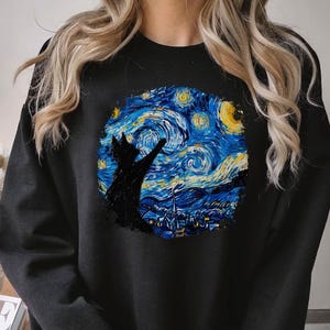 Puede incluir: Sudadera negra con un estampado gráfico de un gato negro mirando hacia arriba a un cielo nocturno estrellado. El cielo está pintado en tonos de azul, amarillo y blanco, que recuerda a la 'Noche estrellada' de Vincent van Gogh.