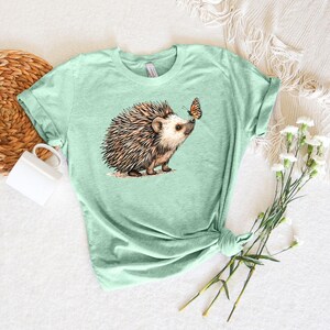 May include: A light green t-shirt with a brown and white hedgehog and a butterfly graphic. The hedgehog is looking at the butterfly.