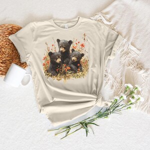 May include: A beige t-shirt with a graphic of three black bear cubs sitting in a field of flowers. The cubs are surrounded by red and orange flowers.