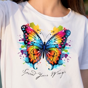 以下が含まれることがあります： カラフルな蝶のグラフィックと「Spread Your Wings」というテキストがプリントされた白いTシャツです。