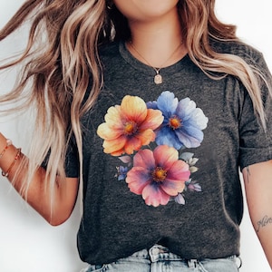 Peut inclure: Un t-shirt gris foncé avec un imprimé floral composé de trois fleurs en aquarelle dans des tons d'orange, de rose et de bleu. Les fleurs sont disposées en grappe et ont un aspect délicat et féminin.
