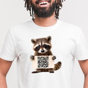May include: White t-shirt featuring a raccoon holding a sign with a QR code. The raccoon is brown and gray with a black mask around its eyes. The QR code is black and white.