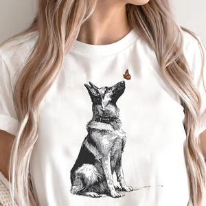 Può includere: T-shirt bianco con un'illustrazione in bianco e nero di un pastore tedesco che guarda una piccola farfalla arancione. Il cane è seduto. La maglietta è realizzata in un materiale morbido.
