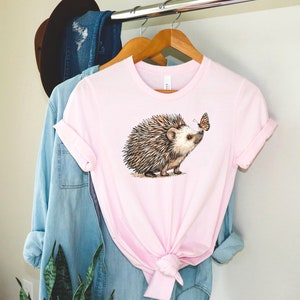 May include: A pink t-shirt with a brown and white hedgehog and a butterfly graphic. The hedgehog is looking at the butterfly.