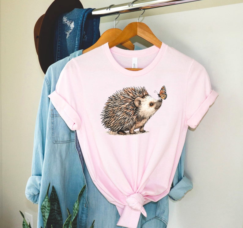 May include: A pink t-shirt with a brown and white hedgehog and a butterfly graphic. The hedgehog is looking at the butterfly.
