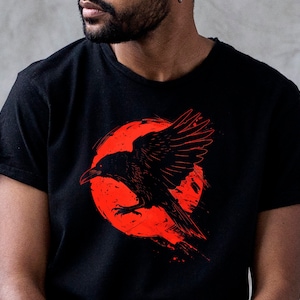 May include: Black t-shirt featuring a striking red and black graphic of a raven in flight against a red circular background. The raven has detailed wing feathers and a piercing gaze. The design has a grunge aesthetic.