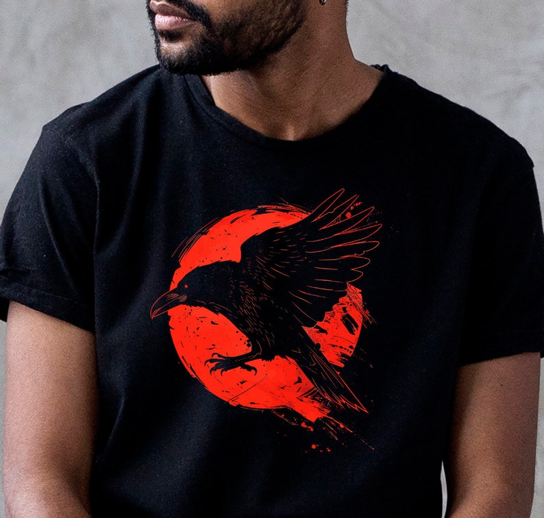 May include: Black t-shirt featuring a striking red and black graphic of a raven in flight against a red circular background. The raven has detailed wing feathers and a piercing gaze. The design has a grunge aesthetic.
