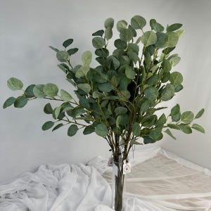 Op de afbeelding: Een boeket van kunstmatige eucalyptusstelen met ronde, groene bladeren. De stelen zijn gerangschikt in een heldere glazen vaas, wat een natuurlijk display creëert. De bladeren variëren in groentinten.