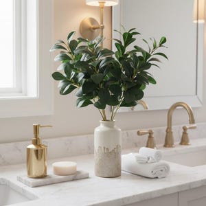 May include: A bathroom countertop arrangement featuring a gold soap dispenser, a marble tray with soap, and a white and beige vase holding green leafy branches. Two rolled white towels are also present, creating a spa-like ambiance.