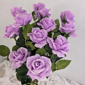 May include: A bunch of lavender roses with green foliage. The roses are in different stages of opening, from buds to fully open blooms. The arrangement is set against a white backdrop.