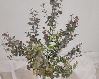 May include: A bunch of artificial eucalyptus stems in a clear glass vase. The leaves are a mix of green, grey, and brown, with a dusty look. The vase has a textured pattern. The arrangement is set against a white background.