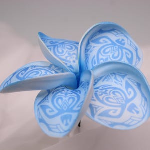 May include: A light blue artificial plumeria flower with white tribal patterns. The flower has five petals, each with a unique design. The petals are slightly curved, giving the flower a realistic appearance. The flower is a decorative item.