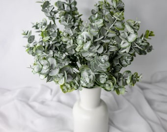 May include: A white ceramic vase displays a bunch of artificial eucalyptus branches. The leaves are a blend of green and grey, with a frosted look. The vase is cylindrical with a rounded top, set against a white fabric background.