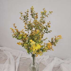 May include: A bouquet of artificial eucalyptus in a clear glass vase. The leaves are a mix of green and yellow, with some tips showing a hint of orange. The vase has a textured design.