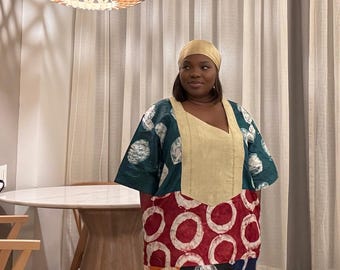 Può includere: Una donna indossa un abito patchwork colorato con corpetto color crema e foulard abbinato. L'abito presenta un mix di fantasie, tra cui cerchi e disegni astratti in vari colori. Una lampada in legno è appesa sopra.