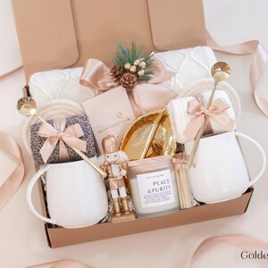 Champagne-toned holiday gift box with white mugs, gold spoons, nutcracker figurine, Peace & Purity candle, cozy socks, and soft cream blanket — a serene and luxurious Christmas gift for couples.