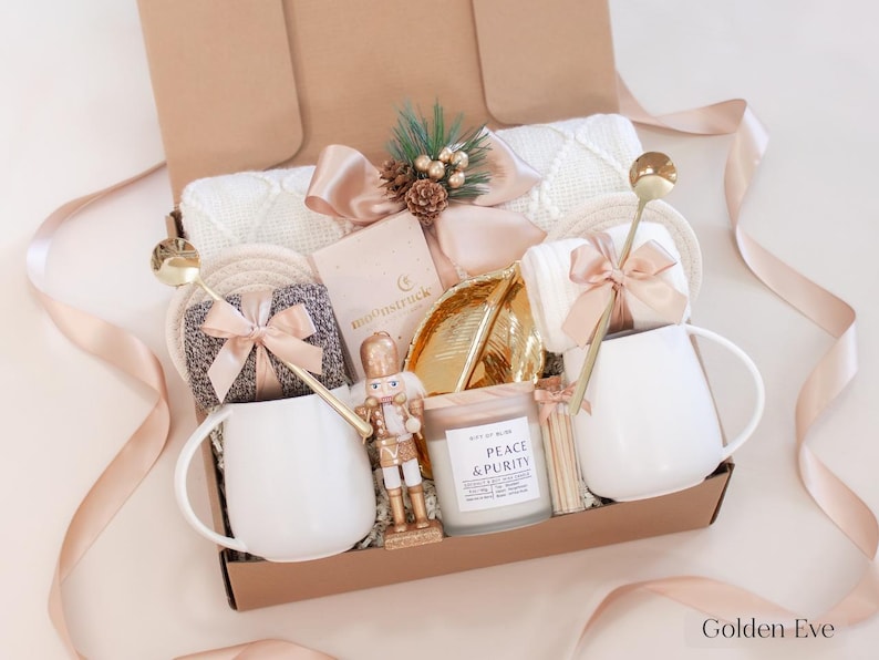 Champagne-toned holiday gift box with white mugs, gold spoons, nutcracker figurine, Peace & Purity candle, cozy socks, and soft cream blanket — a serene and luxurious Christmas gift for couples.