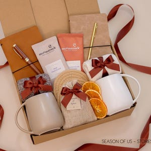 Gift box with journals, luxury wooden pen and gold pen, artisan cocoa, aromatic spice bag for simmer pots or mulling, 2 minimalist mugs, 2 pairs of cozy socks, and 2 cotton yarn coasters, all decorated with satin ribbon in fall color, neatly arranged in a kraft gift box — a thoughtful couples’ gift for reflection and connection.