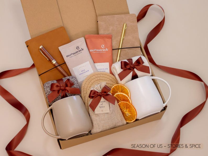 Gift box with journals, luxury wooden pen and gold pen, artisan cocoa, aromatic spice bag for simmer pots or mulling, 2 minimalist mugs, 2 pairs of cozy socks, and 2 cotton yarn coasters, all decorated with satin ribbon in fall color, neatly arranged in a kraft gift box — a thoughtful couples’ gift for reflection and connection.