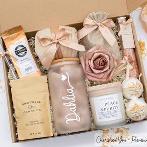 May include: A gift box filled with assorted items, including a coffee bag, a glass jar with "Dahlia" written on it, a candle, a rose, and bath bombs. The box is tied with a ribbon and contains a bag of candy.